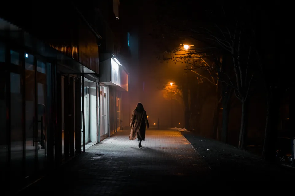 Silhouette of woman in long coat walking on darkened city street alone at night, passing brightly lit sotrefront.